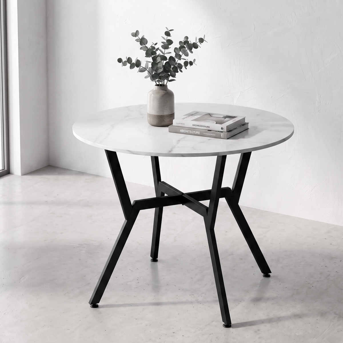 A round dining table with a white marble-patterned top and black metal legs, styled with a vase of greenery and a stack of books in a bright, minimalist room.