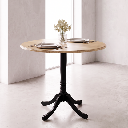 A round wooden dining table with a black pedestal base and three curved legs, styled with plates, silverware, and a small vase of flowers in a bright, modern room.
