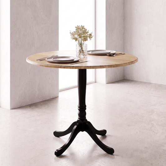 A round wooden dining table with a black pedestal base and three curved legs, styled with plates, silverware, and a small vase of flowers in a bright, modern room.